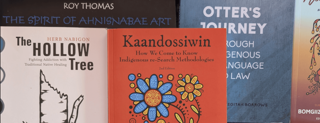 A flat-lay arrangement of Indigenous literature, centered on the red book cover of Kaandossiwin: How We Come to Know: Indigenous re-Search Methodologies (2nd Edition) by Kathleen E. Absolon (Minogiizhigokwe). The cover artwork displays intricate Woodland-style illustrations in red, blue, yellow, and black, depicting stylized floral blooms rising from intertwining figures. Surrounding Kaandossiwin are partially visible covers of related works, including The Hollow Tree by Herb Nabigon, The Spirit of Ahnisnabae Art by Roy Thomas, Otter's Journey Through Indigenous Language and Law by Lindsay Keegitah Borrows, and Anton Treuer's Living Our Language.